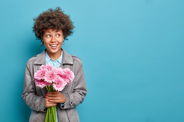 Hermosa mujer alegre sostiene un ramo de gerberas rosas celebra las vacaciones de primavera vestida con un modelo de chaqueta gris contra la pared azul