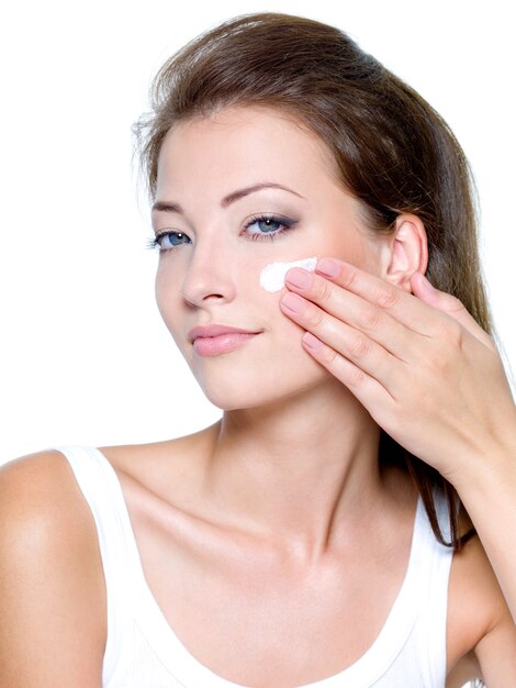 Hermosa mujer aplicando crema cosmética humectante en la cara aislada en blanco