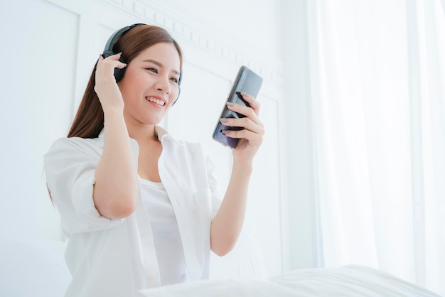 Hermosa mujer asiática atractiva escuchar la canción de la mano de los auriculares sostener el retrato de fondo del dormitorio blanco del teléfono inteligente de mujer asiática de camisa blanca de pelo largo