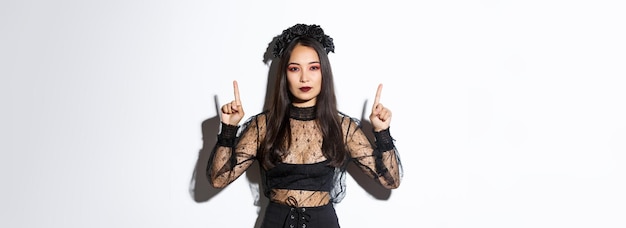 Foto gratuita hermosa mujer asiática atrevida con vestido gótico negro, disfrazada de bruja para halloween y señalando con el dedo hacia arriba, mostrando su logotipo o pancarta en un fondo blanco vacío, fondo blanco