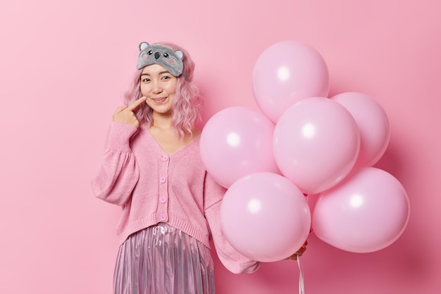 Una hermosa mujer asiática complacida con el pelo teñido de rosa apunta con el dedo en la cara y sonríe agradablemente con un puente de máscara de sueño y poses de falda con globos aislados sobre un fondo rosa. Concepto de fiesta