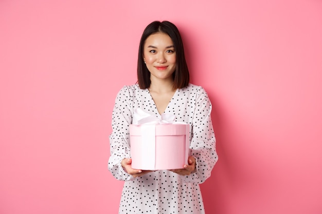 Hermosa mujer asiática deseando felices fiestas dándole un regalo en una linda caja de pie contra el fondo rosa ...