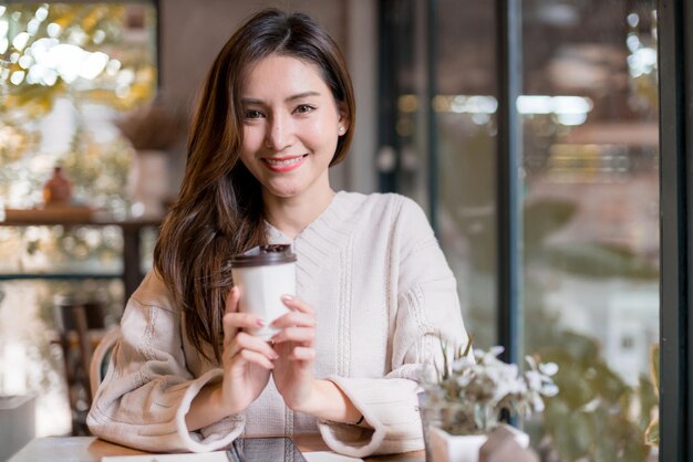 Hermosa mujer asiática disfruta de la bebida caliente por la mañana cerca de la ventana en el concepto de ideas de estilo de vida de la cafetería