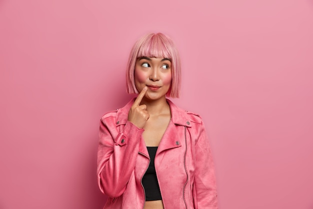 Hermosa mujer asiática de ensueño mira hacia otro lado con expresión pensativa, mantiene el dedo cerca de la boca, vestida con un traje de moda tiene un peinado de moda cabello rosado