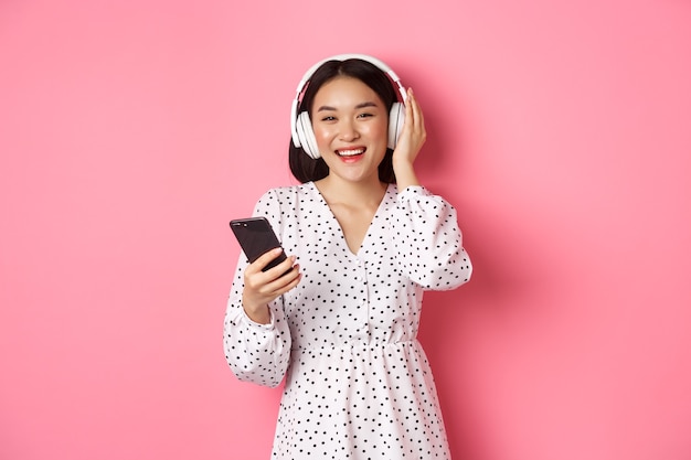 Hermosa mujer asiática escuchando música en auriculares, utilizando teléfono móvil, sonriendo feliz a la cámara, de pie sobre fondo rosa