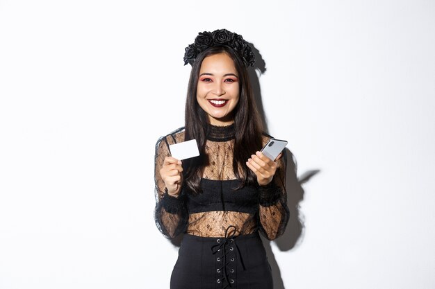 Hermosa mujer asiática con estilo en traje de halloween sosteniendo teléfono móvil con tarjeta de crédito y sonriendo, de pie sobre fondo blanco.