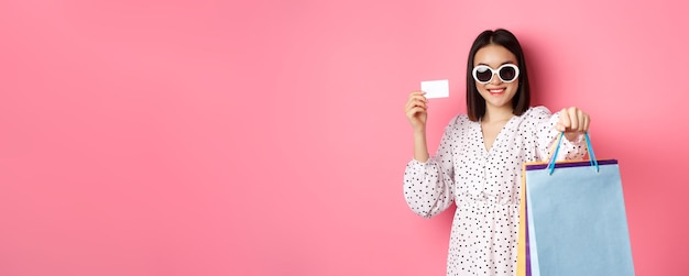 Foto gratuita hermosa mujer asiática con gafas de sol yendo de compras sosteniendo bolsas y mostrando la tarjeta de crédito de pie