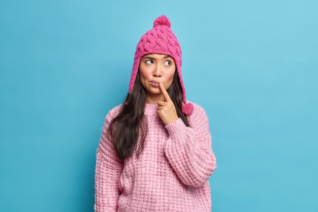 Hermosa mujer asiática joven natural viste un suéter de punto y un sombrero mantiene el dedo cerca de los labios concentrado a un lado piensa en los planes para las vacaciones de invierno aisladas sobre la pared azul