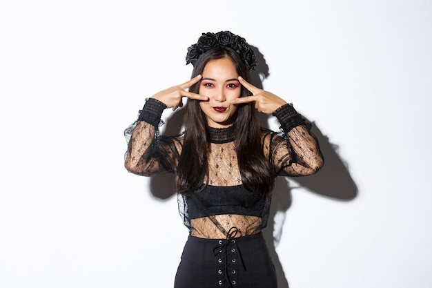 Hermosa mujer asiática con maquillaje gótico, vestido de viuda negra y mostrando gesto de paz, celebrando halloween, de pie sobre fondo blanco.