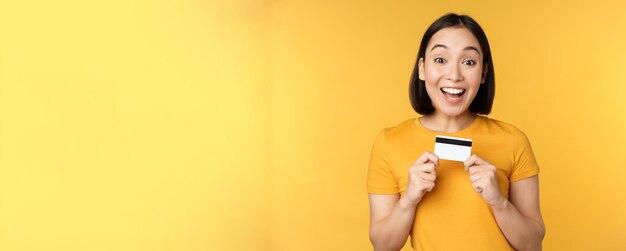 Hermosa mujer asiática mostrando tarjeta de crédito y sonriendo recomendando el servicio bancario de pie sobre fondo amarillo