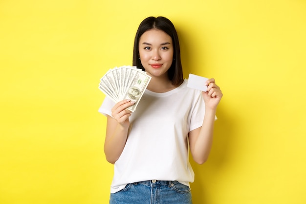 Hermosa mujer asiática con pelo corto y oscuro, vistiendo camiseta blanca, mostrando dinero en dólares y tarjeta de crédito plástica, de pie sobre fondo amarillo.