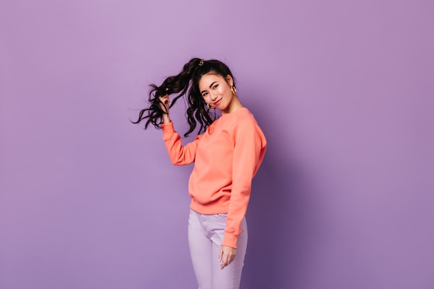 Hermosa mujer asiática en pendientes sobre fondo púrpura. Foto de estudio de la encantadora mujer China rizada jugando con el pelo.