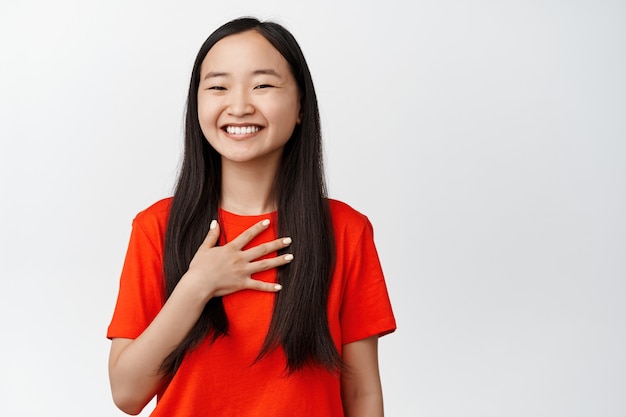 Hermosa mujer asiática riendo y sonriendo, sosteniendo la mano en el pecho, mostrando una emoción natural genuina, de pie en blanco