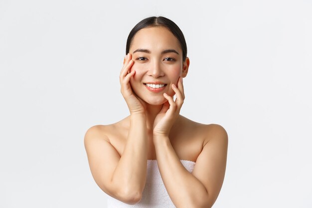 Hermosa mujer asiática sensual en toalla tocando la cara y sonriendo, aplicando productos para el cuidado de la piel, procedimiento cosmético en el salón de spa, masajeando la cara y mirando a cámara feliz, fondo blanco.