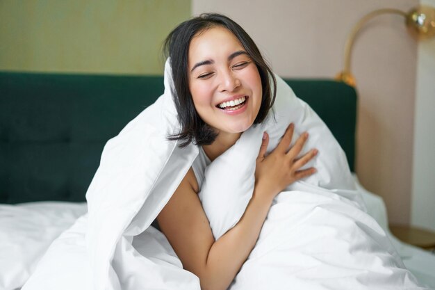 Hermosa mujer asiática sentada en la cama cubierta con edredón blanco sonriendo disfrutando feliz mañana de fin de semana