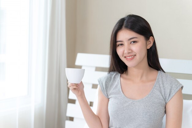 Hermosa mujer asiática sentada en la cama en el dormitorio y sosteniendo la taza de café en la mano con feliz