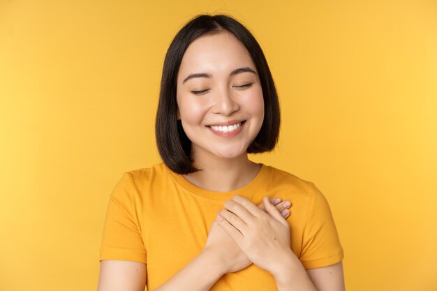 Hermosa mujer asiática sonriendo con ternura y cuidado cogidos de la mano en el corazón de pie en camiseta sobre fondo amarillo