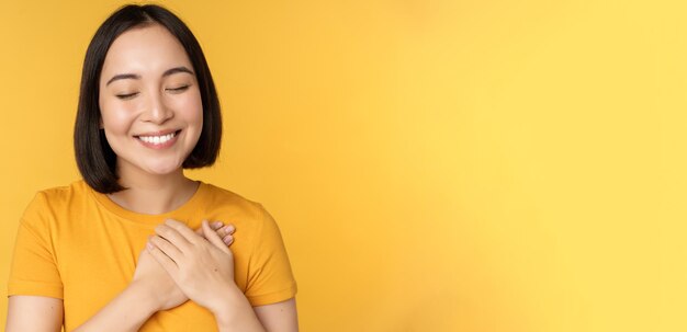 Hermosa mujer asiática sonriendo con ternura y cuidado tomados de la mano en el corazón de pie en camiseta ove