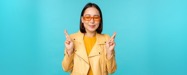 Hermosa mujer asiática sonriente deseando cruzar los dedos para tener buena suerte y luciendo esperanzada de pie sobre un fondo azul