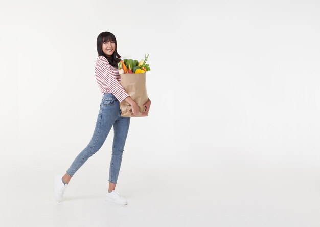 Hermosa mujer asiática sonriente sosteniendo una bolsa de compras llena de comestibles y buscando copiar espacio a un lado