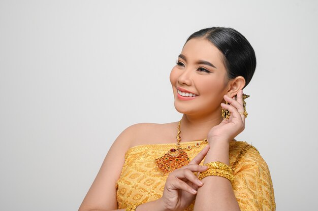 Hermosa mujer asiática en traje tradicional tailandés sonríe y posa con sus aretes con gracia en la pared blanca