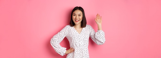 Hermosa mujer asiática vestida diciendo hola agitando la mano para saludar y decir hola sonriendo amablemente cuando llegó