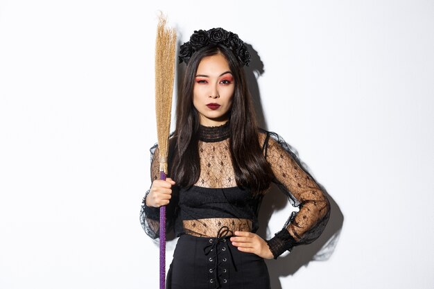 Hermosa mujer asiática en vestido de encaje gótico y corona negra, sosteniendo la escoba y mirando sospechoso a la cámara, de pie sobre fondo blanco.