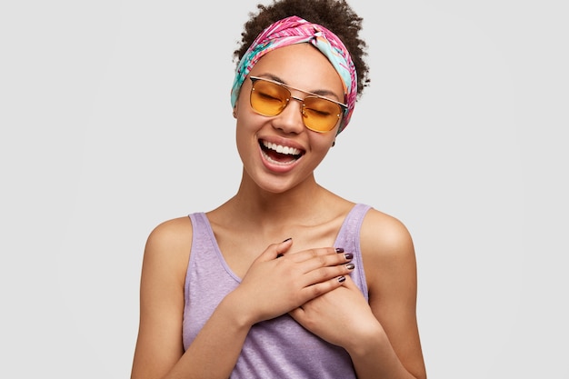 Hermosa mujer de aspecto positivo y amigable mantiene ambas manos en el pecho, expresa gratitud a la hermana por su ayuda y consejo, usa gafas de sol y gafas de sol de moda, aisladas sobre una pared blanca