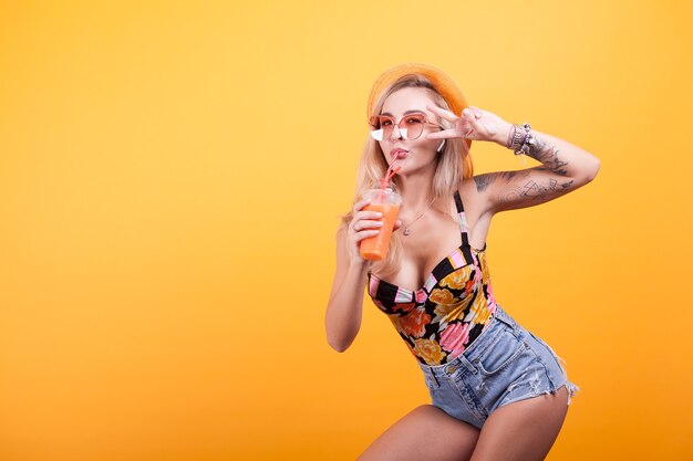 Hermosa mujer atractiva bebiendo jugo de naranja con gafas de sol y sombrero sobre fondo amarillo en estudio