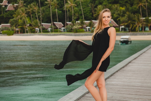 Hermosa mujer atractiva vestida con vestido negro posando en el muelle en el hotel resort de lujo, vacaciones de verano, playa tropical