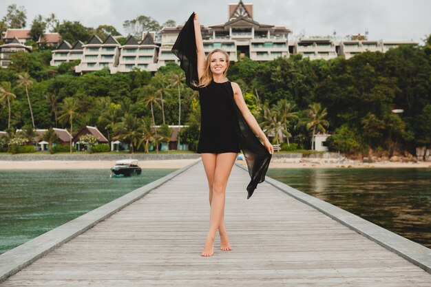 Hermosa mujer atractiva vestida con vestido negro posando en el muelle en el hotel resort de lujo, vacaciones de verano, playa tropical