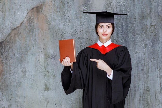 Hermosa mujer en bata de graduación con libro apuntando a algo. foto de alta calidad