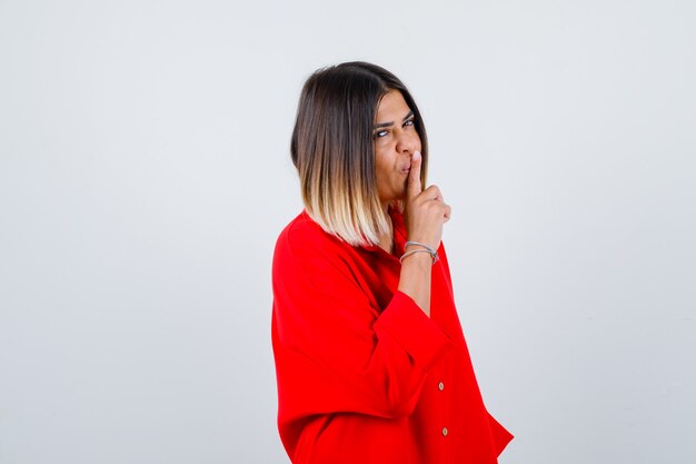 Hermosa mujer en blusa roja mostrando gesto de silencio y mirando sensible, vista frontal.