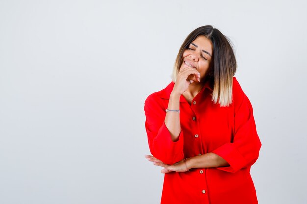 Hermosa mujer en blusa roja sosteniendo la mano en la barbilla, cerrando los ojos y mirando pacífica, vista frontal.