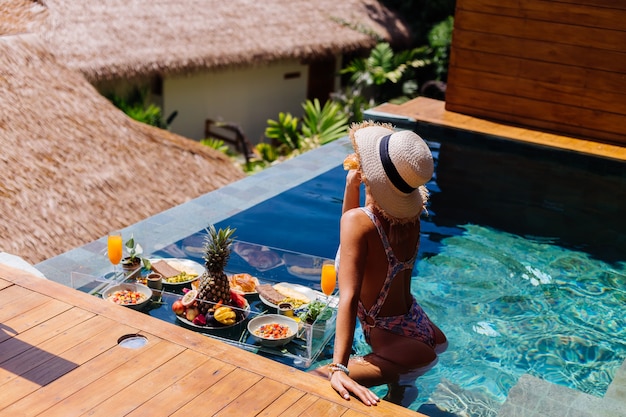 Hermosa mujer bronceada caucásica en bikini y sombrero de paja con desayuno flotante en la increíble villa de lujo de estilo bali en un día soleado junto a la piscina, fondo tropical.