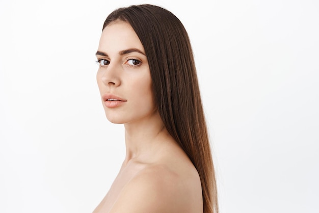 Hermosa mujer con cabello largo y moreno saludable, piel limpia y suave, facial hidratado sin arrugas, efecto antienvejecimiento de los cosméticos, gire la cabeza y luzca fondo blanco de moda