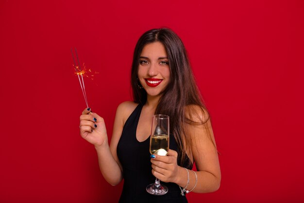 Hermosa mujer con cabello largo oscuro con vestido negro con hombros descubiertos sosteniendo una copa de champán y bengala preparándose para la fiesta de Navidad sobre la pared roja