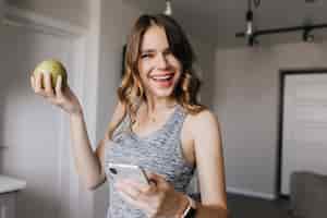 Foto gratuita hermosa mujer con cabello ondulado brillante riendo mientras posa en casa. chica blanca de ensueño con manzana y smartphone.