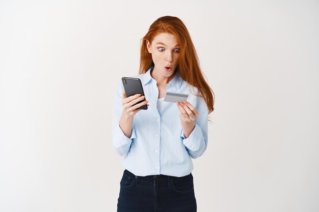 Hermosa mujer con cabello pelirrojo haciendo orden en internet, sosteniendo teléfono inteligente y tarjeta de crédito plástica, pared blanca