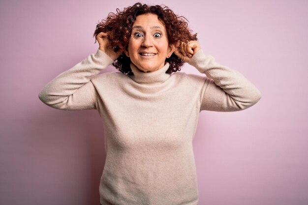 Hermosa mujer de cabello rizado de mediana edad con suéter casual de cuello alto sobre fondo rosa Sonriendo tirando de las orejas con los dedos gesto divertido Problema de audición