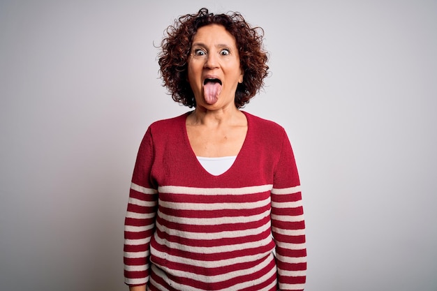 Hermosa mujer de cabello rizado de mediana edad con suéter casual a rayas sobre fondo blanco sacando la lengua feliz con expresión divertida Concepto de emoción