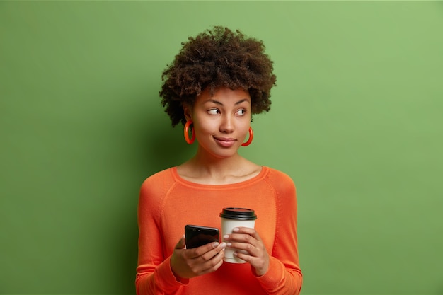 Hermosa mujer con cabello rizado sostiene una taza de papel de delicioso café y el teléfono inteligente usa un suéter naranja casual aislado sobre una pared verde vivo envía mensajes