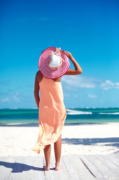 Hermosa mujer caliente en colorido sombrero para el sol y vestido caminando cerca de la playa del océano en un caluroso día de verano en arena blanca