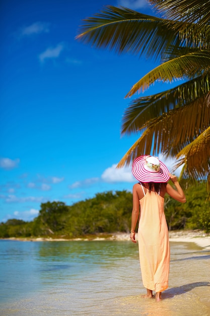 Hermosa mujer caliente en colorido sombrero para el sol y vestido caminando cerca de la playa del océano en un caluroso día de verano cerca de la palma