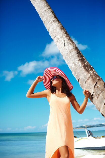 Hermosa mujer caliente en colorido sombrero para el sol y vestido caminando cerca de la playa del océano en un caluroso día de verano cerca de la palma