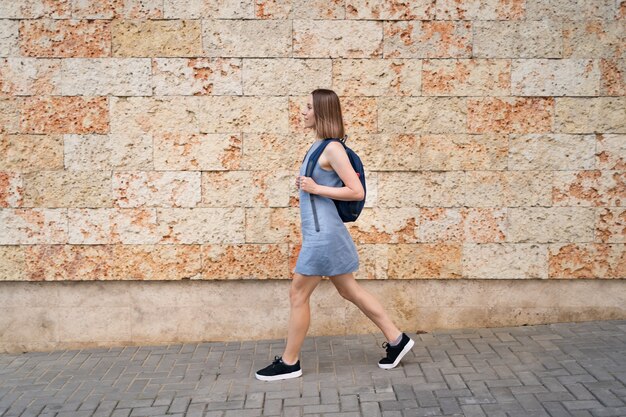 Hermosa mujer caminando con una mochila en vestido azul caminando en la ciudad en la pared decorada con piedra con textura marina