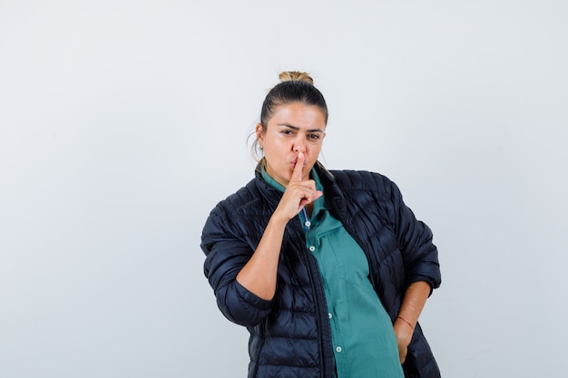 Hermosa mujer en camisa verde, chaqueta negra mostrando gesto de silencio, con la mano en la cadera y mirando seria, vista frontal.