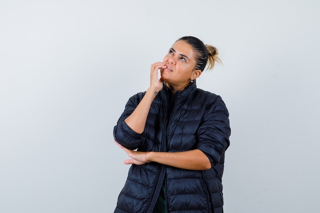 Hermosa mujer en camisa verde, chaqueta negra de pie en pose de pensamiento, poniendo la mano cerca de la boca y mirando pensativo, vista frontal.