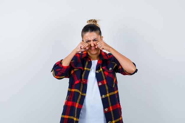 Hermosa mujer en camiseta blanca, camisa cheked con puños cerca de los ojos mientras llora y se ve triste, vista frontal.