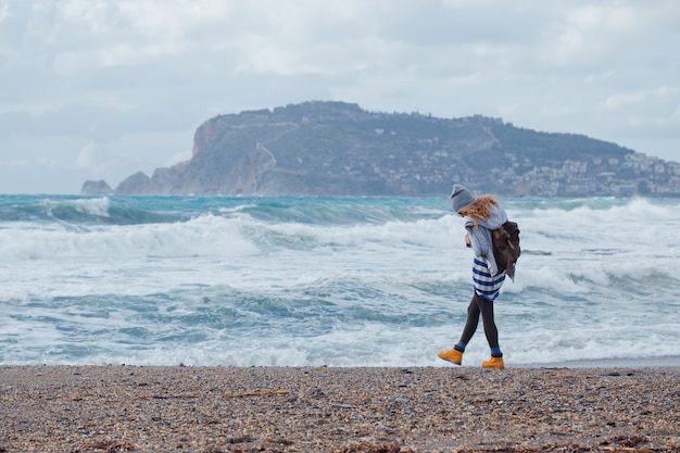 Hermosa mujer con capucha gris y sombrero caminando en la playa durante el día con mar